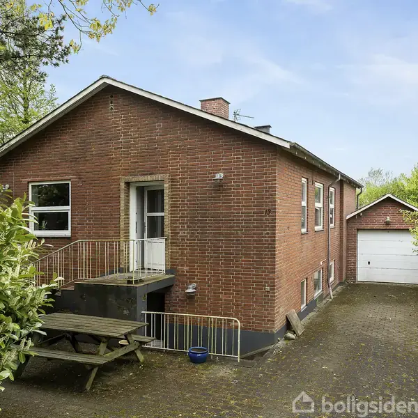 Rødt murstenshus med hvide vinduesrammer og trappe ved indgangen. En bænk står foran huset. Lang indkørsel fører til en garage. Omkringliggende grønne buske og træer.