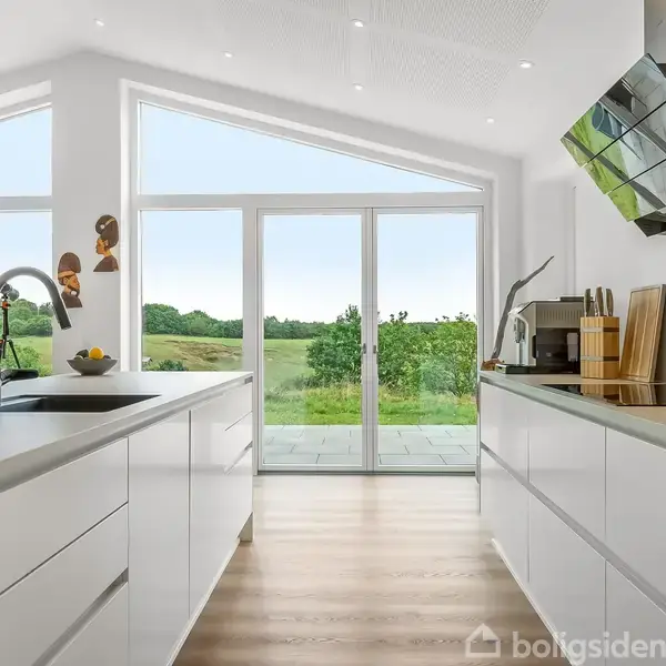 Lyst, moderne køkken med hvide skabe og store vinduer, der åbner udsigten til en grøn have. Et køkkenbord med vask og en stilfuld emhætte er centralt placeret.