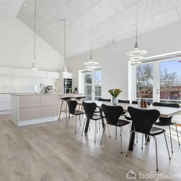Stor spisestue med lang, trædekket bord og sorte stole. Køkkenø i baggrunden. Store vinduer lukker lys ind, planter ses i hjørnet, moderne hængelamper ovenfor. Ingen tekst fundet.