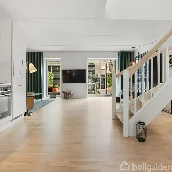 Stue med hvidt køkken til venstre, trappe til højre, der fører til en anden etage, og store glasdøre i baggrunden, der åbner ud til en terrasse med planter.