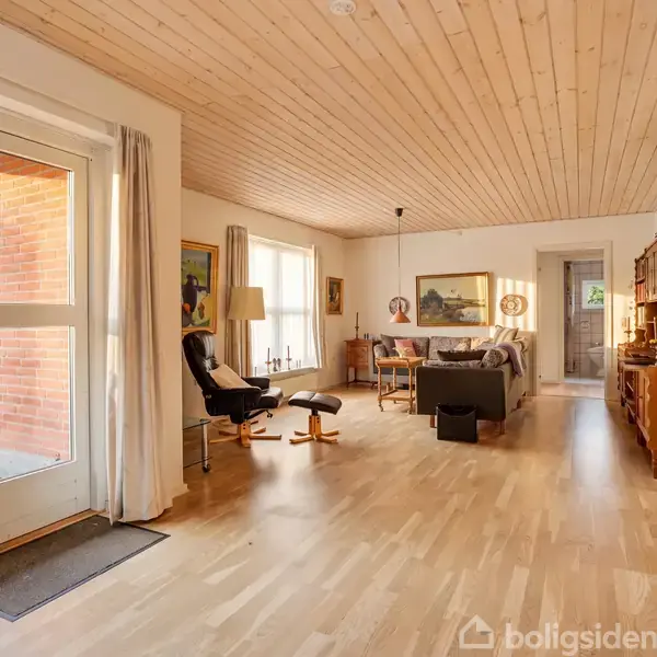 Lænestol og skammel står ved siden af et stort vindue. Sofa og bord i baggrunden, omgivet af træloft og gulv, med bøger på en reol. Belysning fra lamper og dagslys.