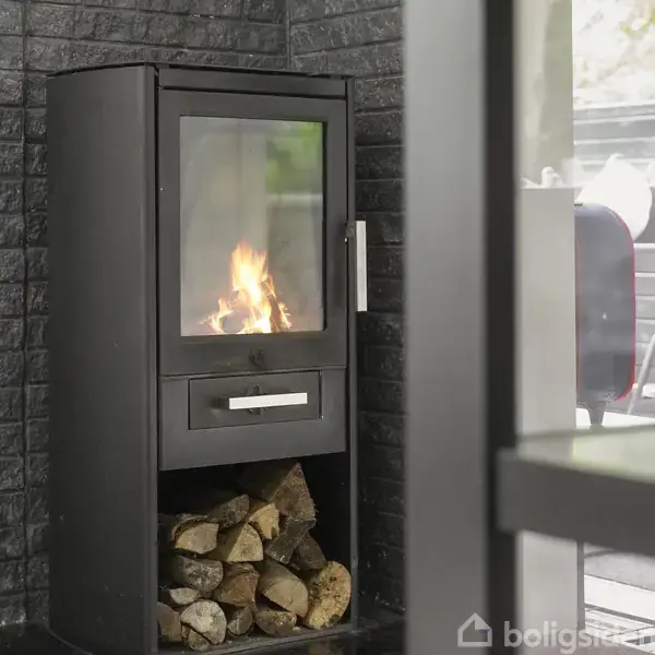 Brændeovn udsender flammer, mens den står i et hjørne af et værelse med mørke murstensvægge. Brændestykker er opbevaret under ovnen, og udenfor ses en have med møbler.