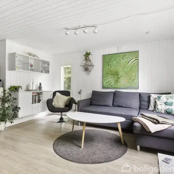 Grå sofa med puder står stille i en lys stue med hvide vægge og træloft. Et rundt bord og en stol står ved siden af, omgivet af planter og vægkunst.