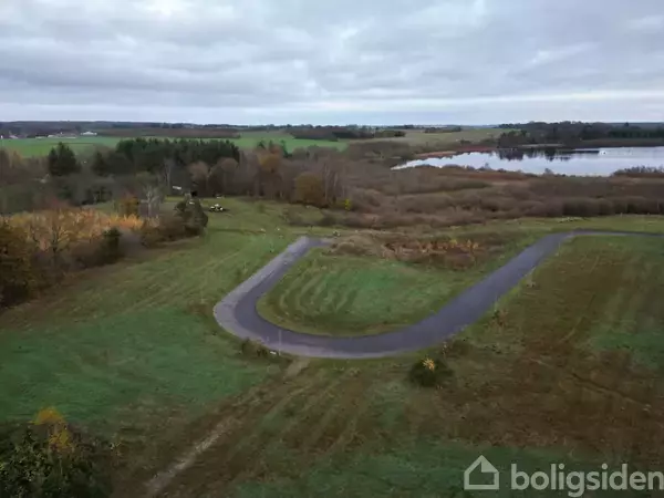 En snoet asfaltvej strækker sig gennem et grønt område med kort græs. Omkringliggende landskab består af skovområder og en sø i baggrunden under en overskyet himmel.