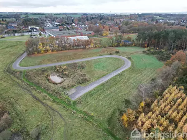 En snoet vej løber gennem et grønt landskab med en lille dam til venstre. Omkringliggende træer har efterårsfarver, og en by anes i horisonten.