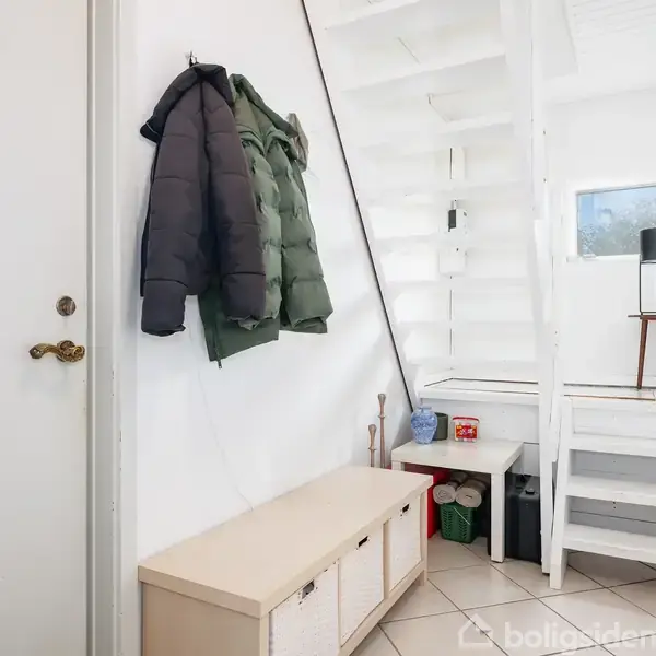 En entre har en hvid trappe, en lille bænk med opbevaring, og to jakker hængende på en krog. Der er også en lampe på et bord ved trappen.