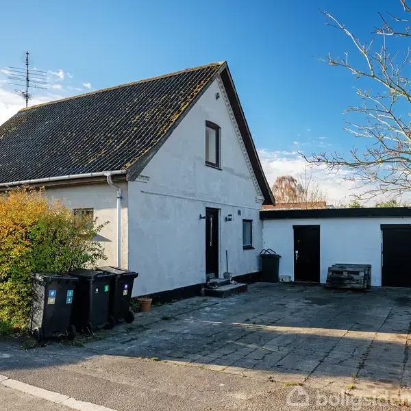 Et hvidt hus med sort tag står ved en flisebelagt indkørsel. Affaldsbeholdere er placeret ved siden af, og en hæk omkranser området. Himlen er blå og træet uden blade.