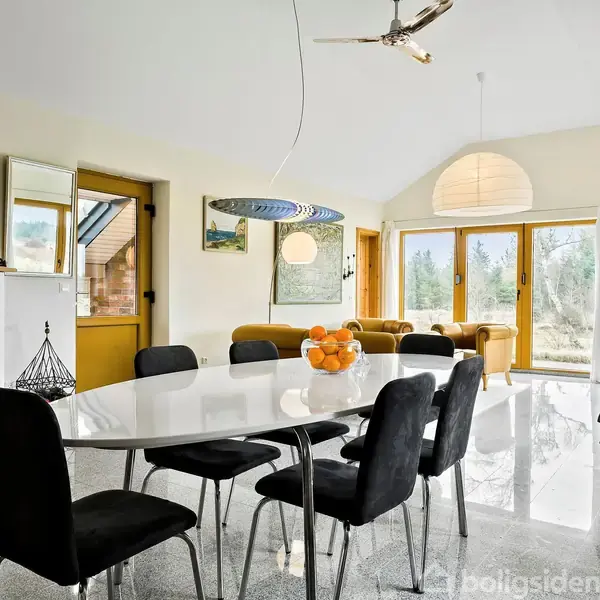 Et ovalt bord med seks sorte stole står i et lyst rum. Bordet er dekoreret med en skål appelsiner. Store vinduer med udsigt til natur omgiver rummet.