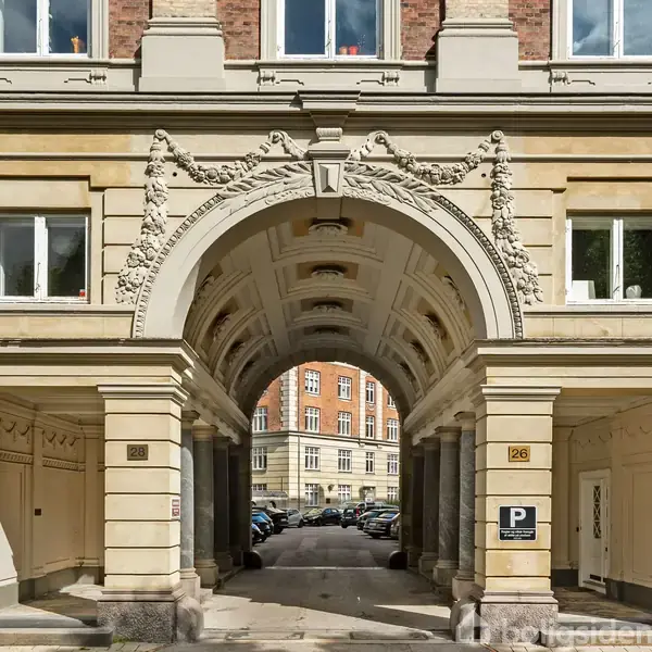 En buegang er dekoreret med udsmykninger, der fører til en indre gårdsplads med parkerede biler. Bygningen omkring har klassiske detaljer og tydelige vinduer. Skilt viser "P" for parkering.