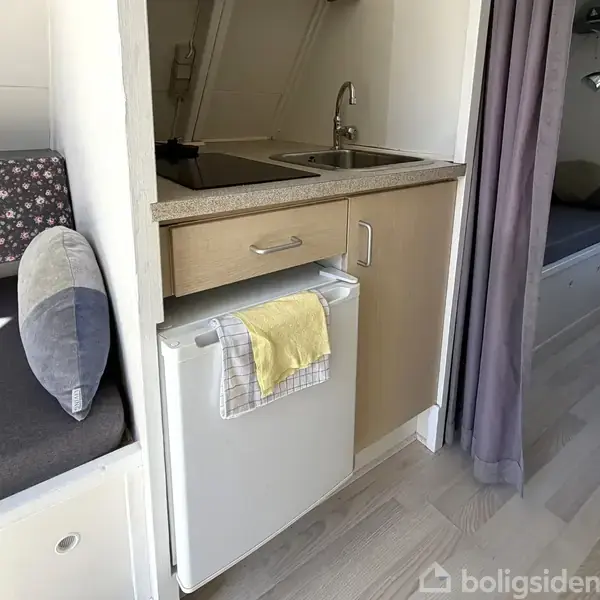 En lille køkkenkrog med håndvask og kogeplade er omgivet af en seng og sofa. Et håndklæde hænger på et lille køleskab. Rum med lyst trægulv og enkel indretning.