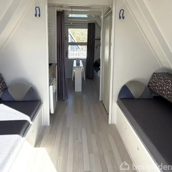 To senge er placeret på hver side af en smal gang. Lyst trælaminatgulv og hvide vægge skaber et lyst rum. En dør fører til et andet rum med vindue.