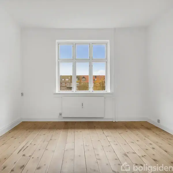 Et tomt rum med lyse vægge og trægulv. Et stort vindue giver udsigt til farverige bygninger under en blå himmel. En radiator er monteret under vinduet.