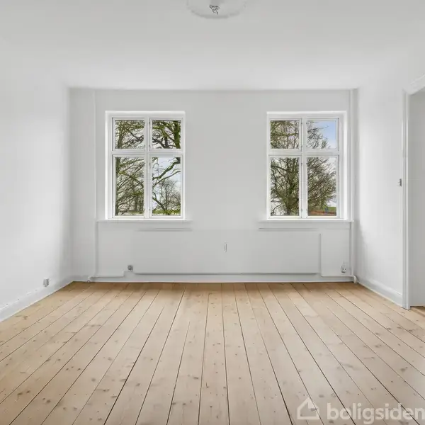 Et tomt værelse med trægulv har to store vinduer, der vender mod grønne træer udenfor. Væggene er hvide, og der er en radiator under vinduerne.