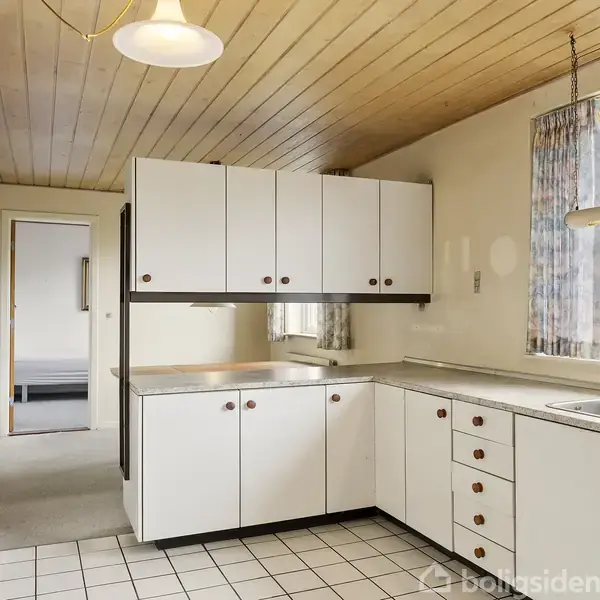 Køkken med hvide skabe og skuffer ved køkkenvasken, flisegulv og træloft. En gang fører til et andet rum med seng. Vinduer har blomstrede gardiner og udsigt til have.