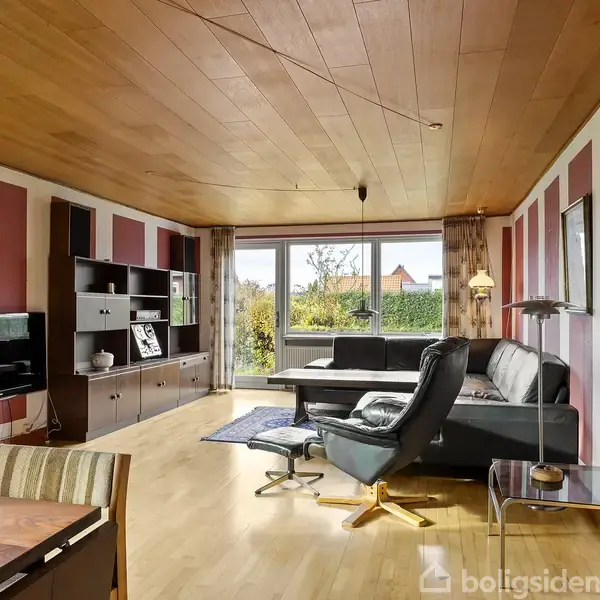 Stue med en sort lædersofa, placeret ved et stort vindue med udsigt til en have. Der er reoler langs væggen, et fladskærms-tv og trægulv samt loft.