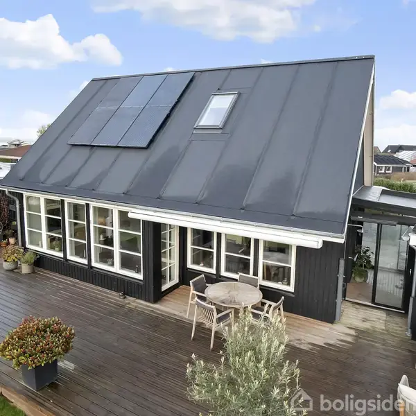 Et sortmalet hus med skråtag har solpaneler på taget. Foran huset, en træterrasse med borde, stole og plantekrukker. I baggrunden ses en parcelhusklynge under en blå himmel.