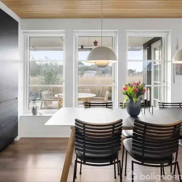 Et moderne spisebord med sorte stole står i et lyst rum. Bordet har en vase med tulipaner. Store vinduer giver udsigt til en terrasse med udemøbler. To hængelamper hænger over bordet.