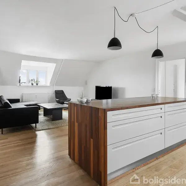 Køkkenø med hvidt skab og træbordplade dominerer rummet, omgivet af en stue med sort sofa og lænestol ved et skråvindue. Hængelamper hænger over øen.