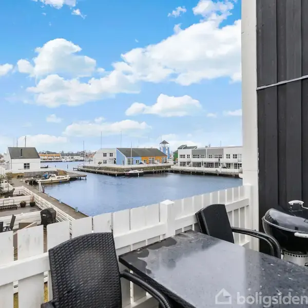 Bord og stole står på en altan med udsigt over en marina. Bygninger og både ses i baggrunden under en blå himmel med skyer.