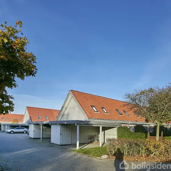 Rækkehusene med røde tegltage står langs en stille vej i et boligkvarter. Træer med efterårsblade omkranser området, og der ses en gadelampe ved siden af indkørslen.