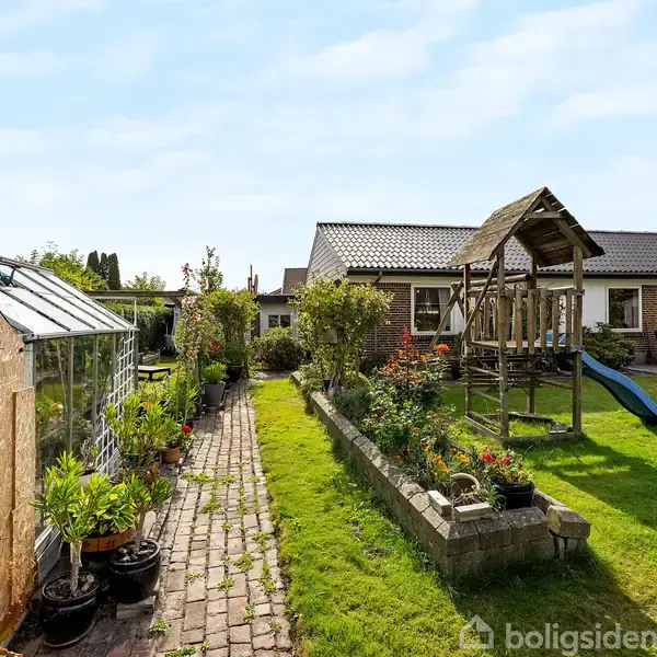 Drivhus med planter langs en sti. I baggrunden ses en rutsjebane og klatrestativ i en have med græs og blomsterbede, foran et hus med en flisebelagt terrasse.