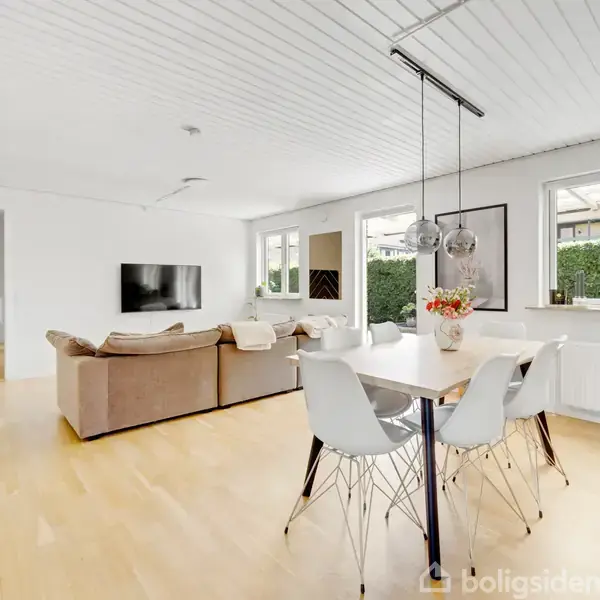 Stuen indeholder en brun sofa og et spisebord med hvide stole. Store vinduer giver lys og udsigt til haven. Lyse trægulve og hvide vægge skaber en moderne atmosfære.