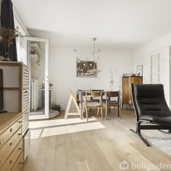 Stue med sort lænestol, placeret på tæppe, foran fladskærms-tv på trækommode. Spisebord med stole står ved åbent glasdør, der giver udsigt til solbelyst veranda.