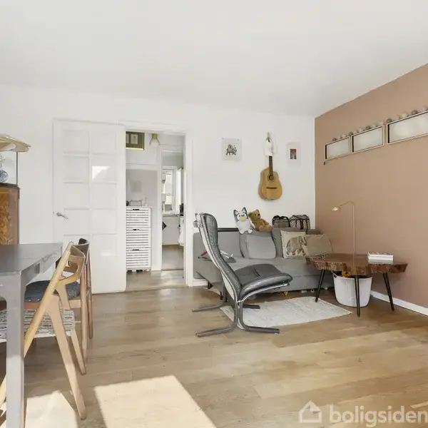 Et lyst stueområde med spisebord og stole til venstre, en lænestol og lille bord til højre. En guitar hænger på væggen. Trægulv og delvis pink væg.