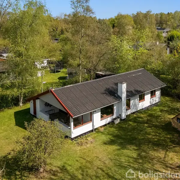 Et enkelt, hvidt hus med sort tag står stille i en grøn have, omgivet af træer og andre små huse i et fredfyldt område.