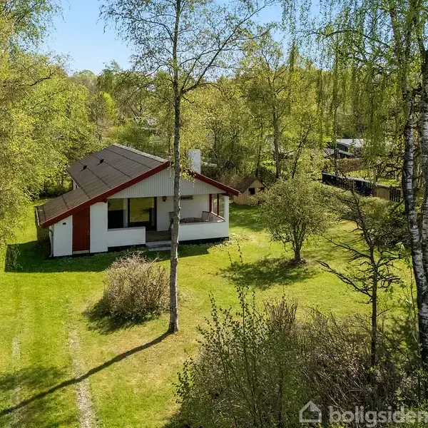 En enkeltstående hus beliggende i en grøn have omkranset af træer. Huset har en brun tag og en hvid facade med røde detaljer. Omkringliggende natur danner en stille skovamnidsætning.