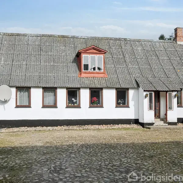 En hvidmalet landbygning med røde døre viser flere små vinduer og en kvist med tagvindue. Bygningen er omgivet af en gårdsplads med brosten og en klar himmel i baggrunden.