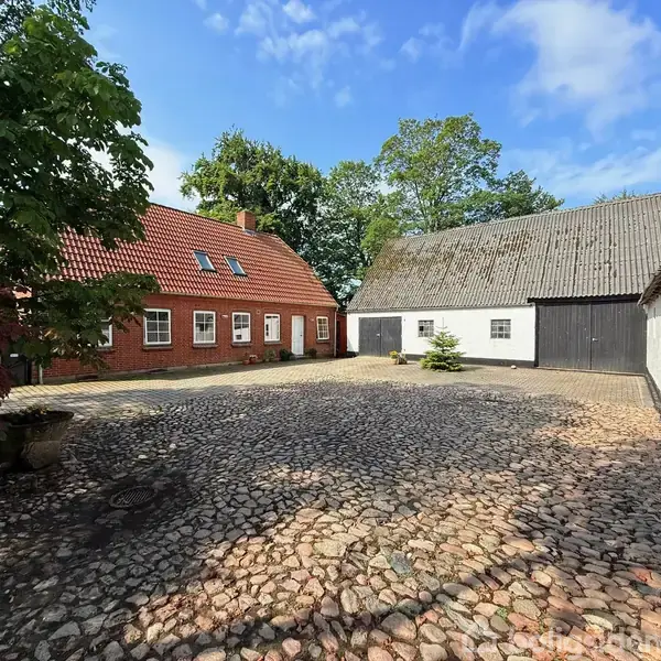 Et rødt murstenshus med rødt tegltag og hvide vinduer står i en gårdsplads med brosten. Til højre ses en hvid og sort ladebygning, omgivet af træer og blå himmel.