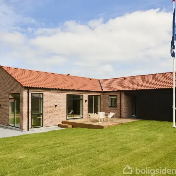 En moderne bungalow med rødt tegltag og store vinduer står på en velplejet græsplæne. Foran huset er der en træterrasse med stole, og en flagstang ses i baggrunden.