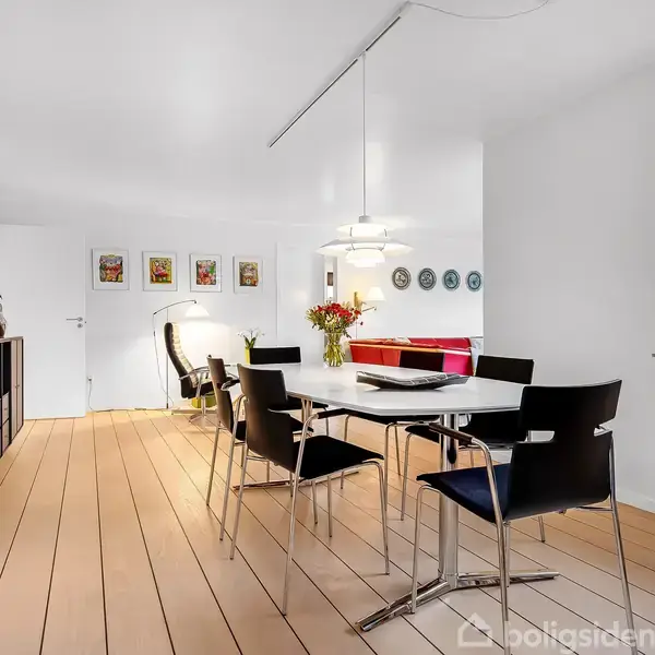 Et moderne spisestueområde har et bord med seks stole, ovenpå hænger en lampe. Til venstre er et sort skab med planter. Væggene har billeder og dekorationer.