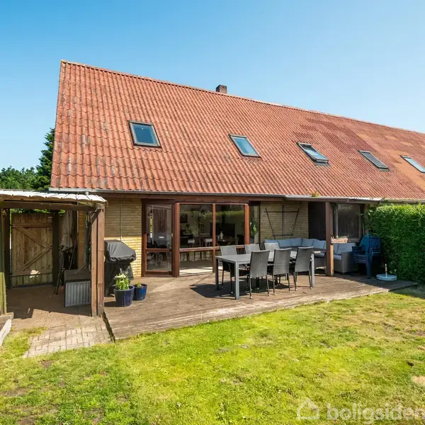 Rækkehus med rød tegltag og store vinduer vender mod en solrig terrasse med havemøbler. Omgivet af en grøn græsplæne og en hæk. Ingen tekst fundet.