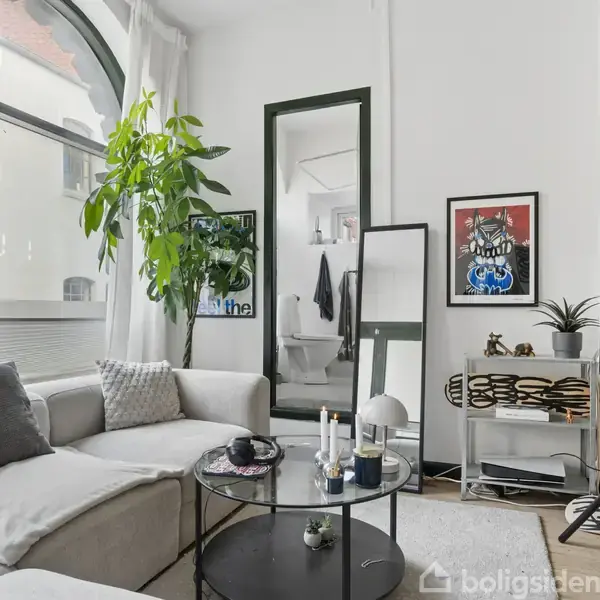 En lys stue med en stor, blød sofa til venstre og et tv til højre. Et rundt glasbord står i midten med stearinlys. Et stort vindue og planter er i baggrunden.