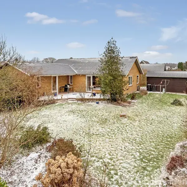 En gul murstensvilla i et villakvarter, med let snedækket have omgivet af buske og træer. I baggrunden ses flere huse og en blå himmel med få skyer.