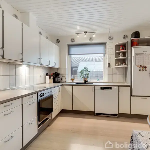 Et lyst køkken med hvide skabe og apparater. En ovn og opvaskemaskine er indbygget, mens køleskabet har magneter. Et bord med stole står til højre. Vindue med plante udenfor.