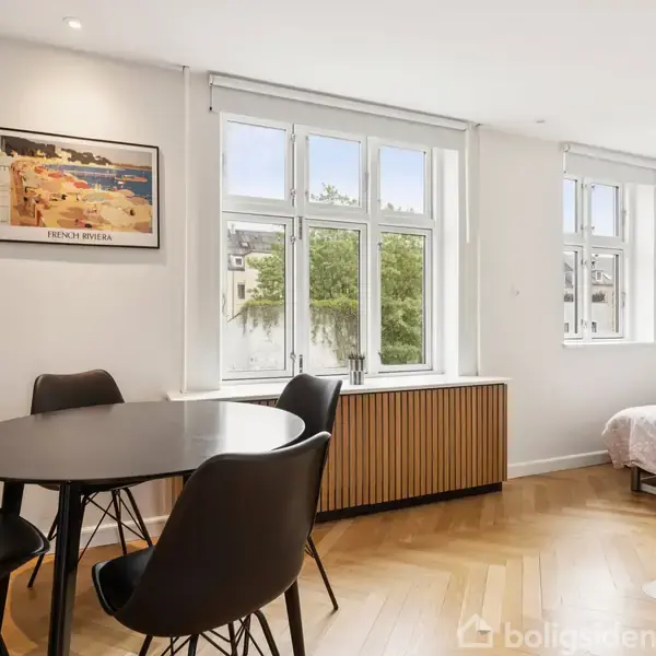 Et rundt, sort spisebord med tre stole står i et lyst rum med parketgulv. Et vindue og et billede på væggen skaber en indbydende atmosfære.