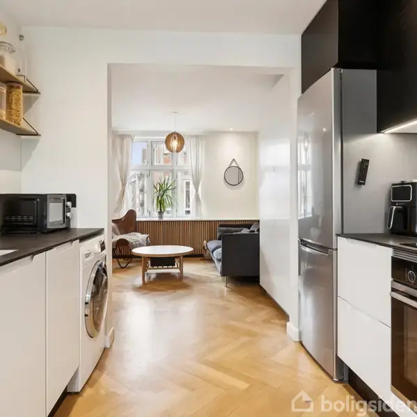 Et moderne køkken med hvide skabe og en stålovn ved siden af køleskabet. Der er hylder med glas og krukker. I baggrunden er der en stue med sofa og vindue.