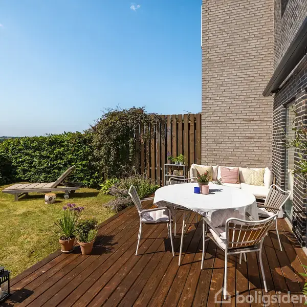 Terrasse med spisebord dækket af en hvid dug, omgivet af stole. Et græsareal med liggestol og planter indrammer scenen. Udsigt over et landskab og under en klar, blå himmel.