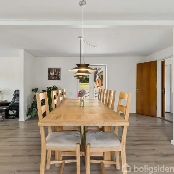 Træbord med otte stole står i en rummelig spisestue. En moderne lampe hænger over bordet. Stuen har lyse vægge, trægulv og synlige døre i baggrunden.