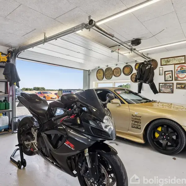 En sort motorcykel står i en garage ved siden af en guldfarvet sportsvogn. Væggene er udsmykket med plakater og hjul, og der er åbne hylder med værktøj og olieprodukter.