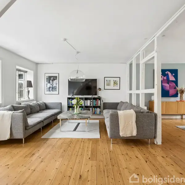 Grå sofaer står omkring et sofabord i en stue med trægulv. Store vinduer og kunst på væggene skaber et lyst miljø. Fjernsynet er placeret på en hylde.