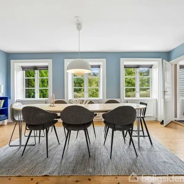 Spisebord med seks stole står på et gråt tæppe i et lyst rum med tre store vinduer. En pendellampe hænger over bordet, og der er opbevaringsmøbler i baggrunden.