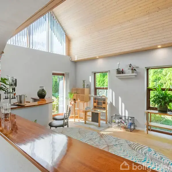 En moderne stue med træloft, store vinduer og grønne planter. Et rundt bord med stole står ved vinduet, og reoler pryder væggen med bøger og dekorationer.