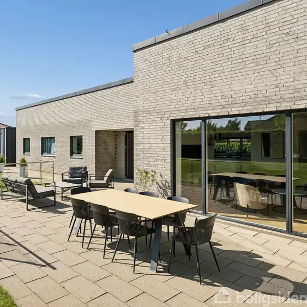 En moderne terrasse med bord og stole foran et hus med store glasvinduer. Terrassen ligger i en velholdt have med grønne græsplæner og buske.