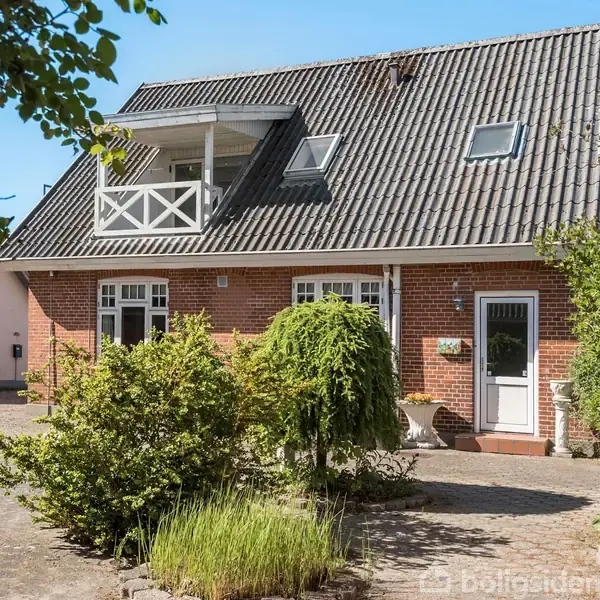 Et rødt murstenshus med grå tagsten har en hvid dør og vinduer. En altan er synlig på taget. Forhaven har grønne buske og planter, omgivet af en gårdsplads med grus.