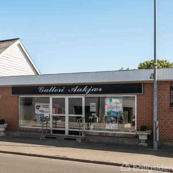 Butiksfacade viser "Galleri Aakjær" på et skilt over en murstensbygning. Store glasvinduer afslører et kunstgalleri indeni. Fortov udenfor, klar blå himmel over.