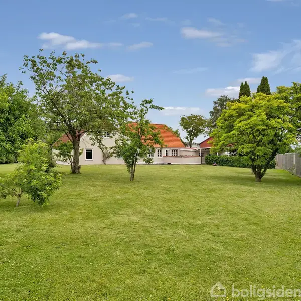 En velholdt græsplæne strækker sig foran et hus med rødt tag og hvide vægge. Træer står spredt på plænen, omkranset af en blå himmel og enkelte skyer.